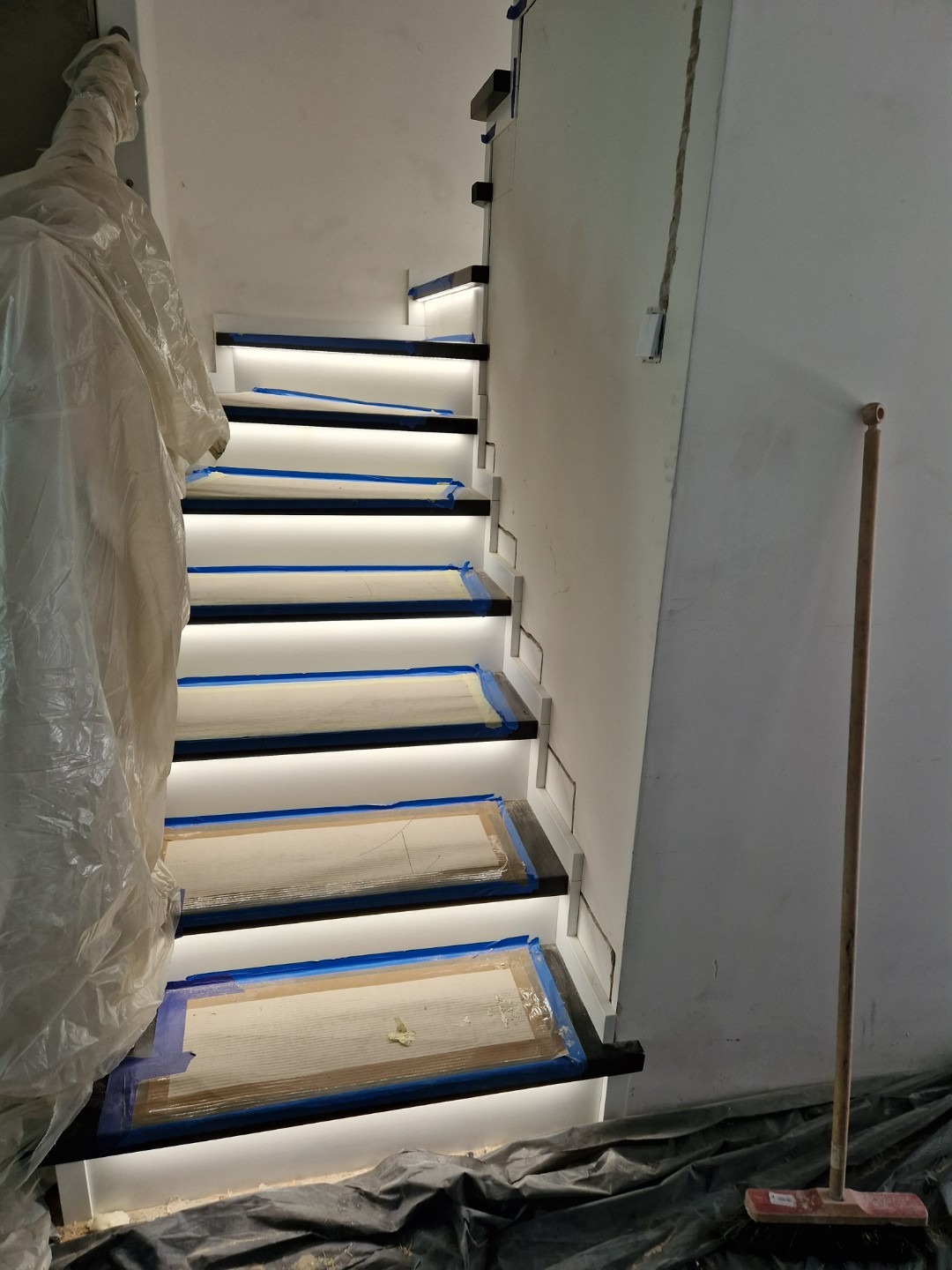 Montaż podświetlanych schodów z czarnymi stopnicami, zabezpieczonymi taśmą malarską i kartonem, widoczna biała ściana, przykryte folią podłoże i szczotka.