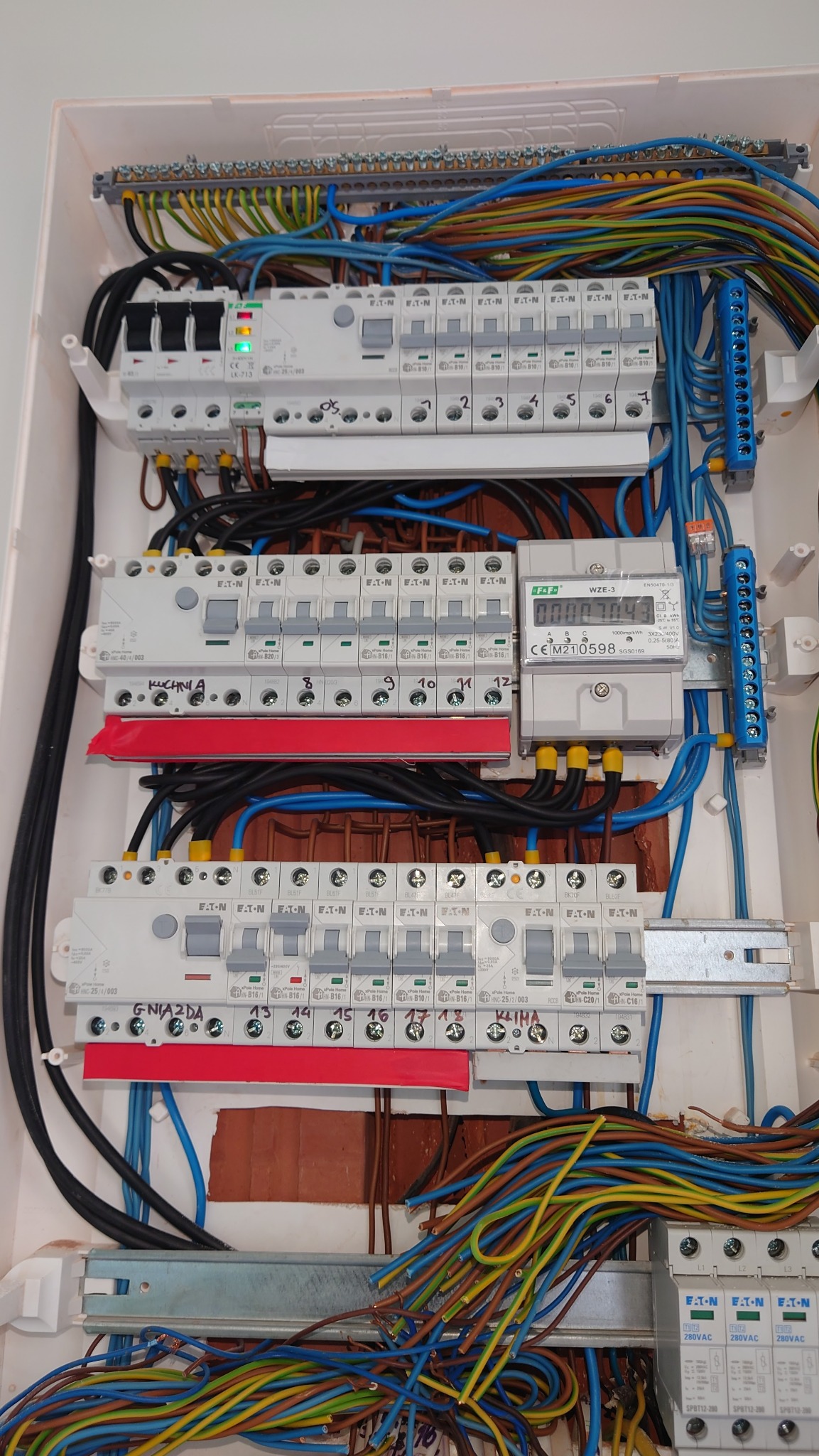 Otwarta skrzynka rozdzielcza z bezpiecznikami, licznikiem energii i licznymi przewodami w różnych kolorach, widoczne oznaczenia obwodów 'Kuchnia' i 'Klima' na naklejkach.