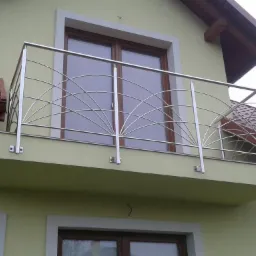 Balkon z ozdobną, nierdzewną balustradą o motywie wachlarza, zamontowaną na elewacji domu o jasnej kolorystyce.