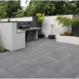Nowoczesny ogród z szarymi płytami tarasowymi, zabudowanym grillem z kamiennym blatem, minimalistyczną kaskadą wodną na tle ściany z szarego kamienia oraz roślinami ozdobnymi w donicach i rabatach.