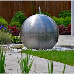 Metalowa kula z fontanną na szczycie, otoczona białymi kamieniami, wkomponowana w zadbany ogród z kostką brukową i różnorodną roślinnością.