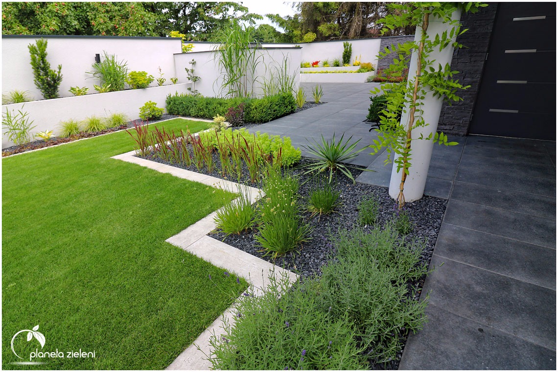 Nowoczesny ogród z geometrycznymi rabatami, równo przystrzyżonym trawnikiem, białym murem w tle i elementami dekoracyjnymi z kamienia.