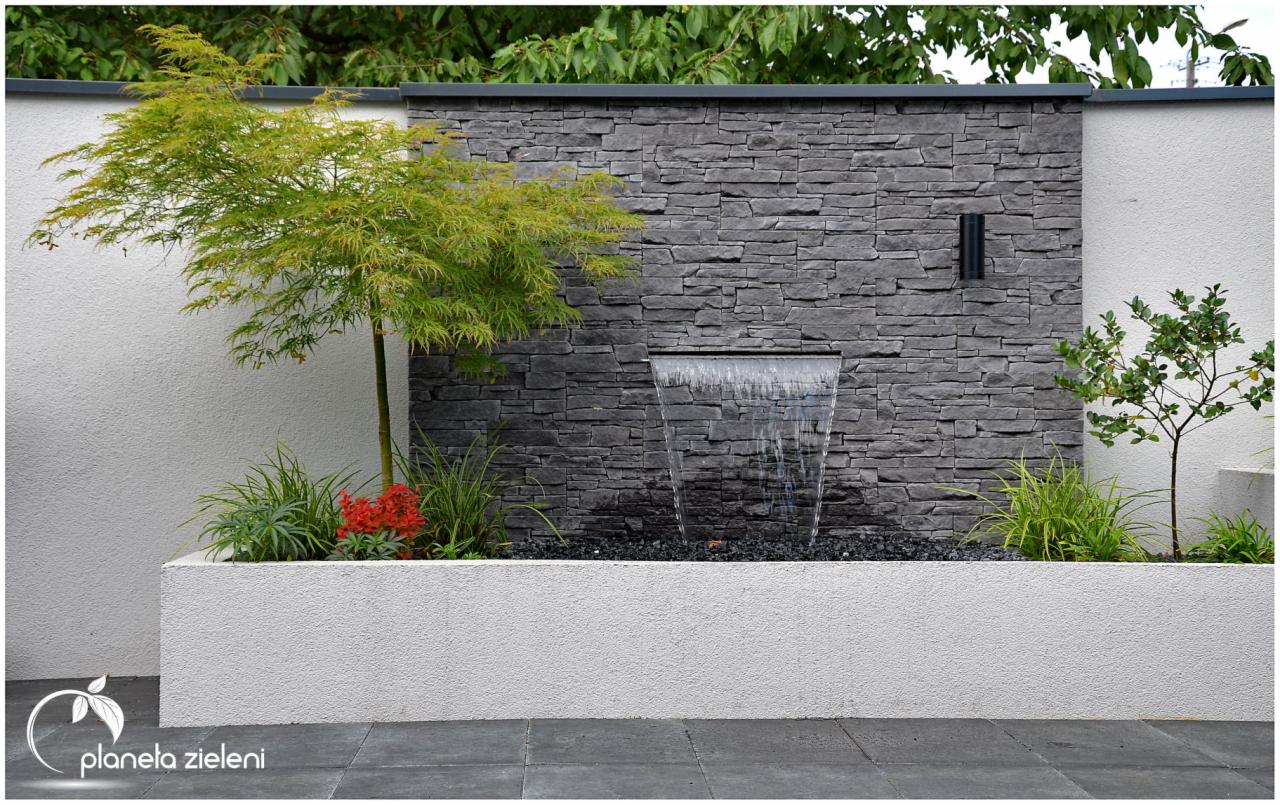 Nowoczesny ogród z kaskadową fontanną na tle ściany z szarego kamienia łupanego, otoczony zielenią i małym drzewkiem z delikatnymi liśćmi.