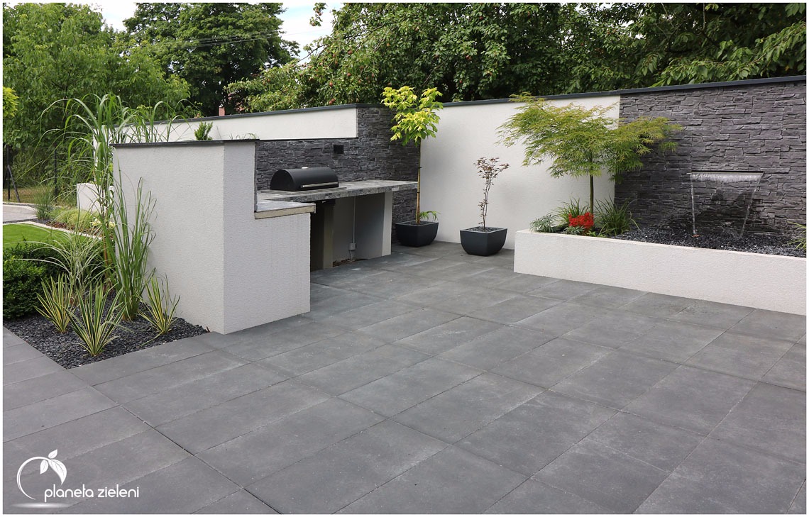 Nowoczesny ogród z szarymi płytami tarasowymi, zabudowanym grillem z kamiennym blatem, minimalistyczną kaskadą wodną na tle ściany z szarego kamienia oraz roślinami ozdobnymi w donicach i rabatach.