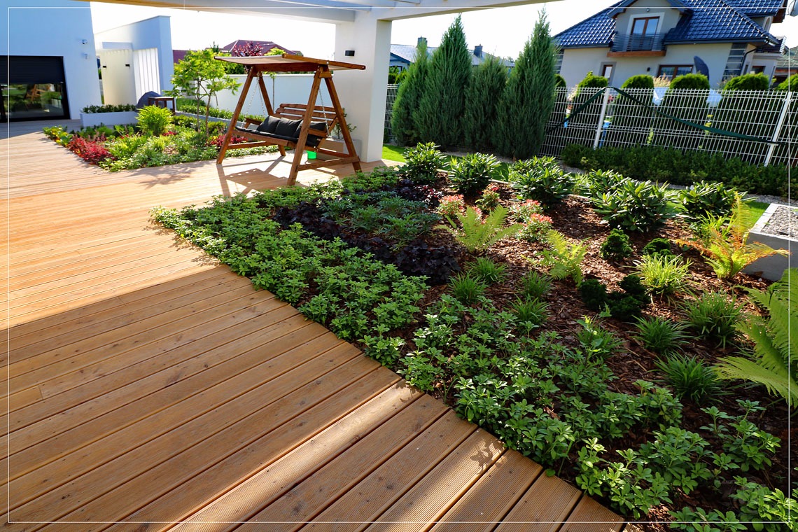 Nowoczesny ogród z drewnianym tarasem i huśtawką pod zadaszeniem, rabaty z różnorodną roślinnością, w tle dom z ogrodem.