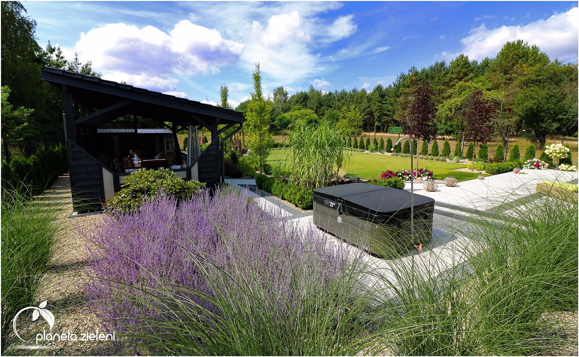 Nowoczesny ogród z ciemną altaną, jacuzzi z pokrywą i kwitnącymi lawendami na pierwszym planie, w tle zadbany trawnik i drzewa.