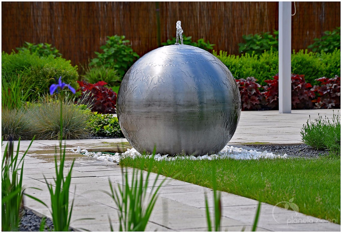 Metalowa kula z fontanną na szczycie, otoczona białymi kamieniami, wkomponowana w zadbany ogród z kostką brukową i różnorodną roślinnością.