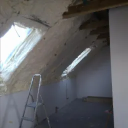 Poddasze po izolacji termicznej, widoczne okna dachowe i metalowa drabina oparta o ścianę, surowe wykończenie wnętrza.