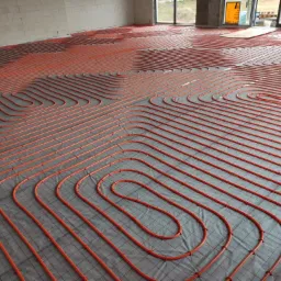 Pomarańczowe rury ogrzewania podłogowego ułożone spiralnie na szarej macie, widok z góry na dużą powierzchnię w surowym stanie.