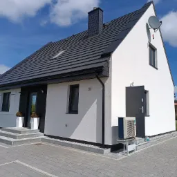 Nowoczesny dom jednorodzinny z zainstalowaną zewnętrzną jednostką klimatyzacji, widoczną przy ścianie budynku obok drzwi wejściowych. Dach pokryty ciemną dachówką, elewacja biała, kostka brukowa...