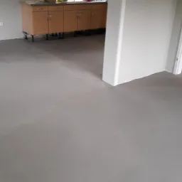 Wyrównana, szara wylewka samopoziomująca na podłodze w pomieszczeniu przed dalszymi pracami wykończeniowymi, widoczne ślady po rozprowadzaniu masy, w tle szafki kuchenne.