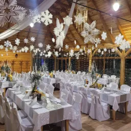 Uroczysta sala z drewnianą konstrukcją, udekorowana białymi płatkami śniegu i elegancko nakrytymi stołami, gotowa na przyjęcie świąteczne lub zimowe.