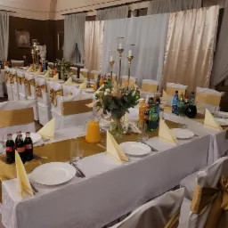 Uroczysty st&oacute;ł z białym obrusem, złotymi bieżnikami i dekoracjami. Krzesła z kokardami, napoje, zastawa i kwiaty. Przygotowania do imprezy.