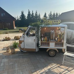 SMAKPOL-CATERING sp. z o.o. - Mobilny bar prosecco 'Bella Ciao' na zewnątrz, z neonowym napisem, kwiatami i butelkami. Drewniany domek z nalewakiem i szklankami na kostce brukowej.