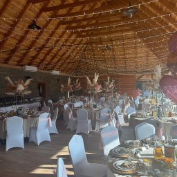 SMAKPOL-CATERING sp. z o.o. - Elegancka sala bankietowa z drewnianym sufitem, udekorowana girlandami świetlnymi i kwiatowymi kompozycjami na stołach z białymi krzesłami i różowymi akcentami.