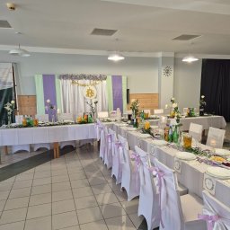 SMAKPOL-CATERING sp. z o.o. - Uroczysty stół komunijny z dekoracją kwiatową, białymi obrusami i krzesłami z fioletowymi wstążkami. Napoje i dania na stole. Pastelowe tło z napisem IHS.