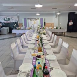 SMAKPOL-CATERING sp. z o.o. - Uroczysty, długi stół z białymi krzesłami, nakryty do przyjęcia. Dekoracje kwiatowe, napoje i zastawa. W tle baner cateringowy i pracownik w uniformie.