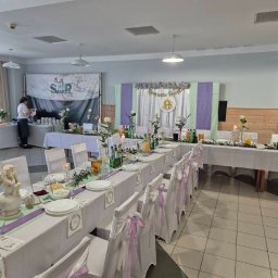 SMAKPOL-CATERING sp. z o.o. - Uroczysty stół komunijny: białe obrusy, pastelowe wstążki na krzesłach, kwiaty, świece, napoje. W tle baner cateringowy i dekoracja IHS.