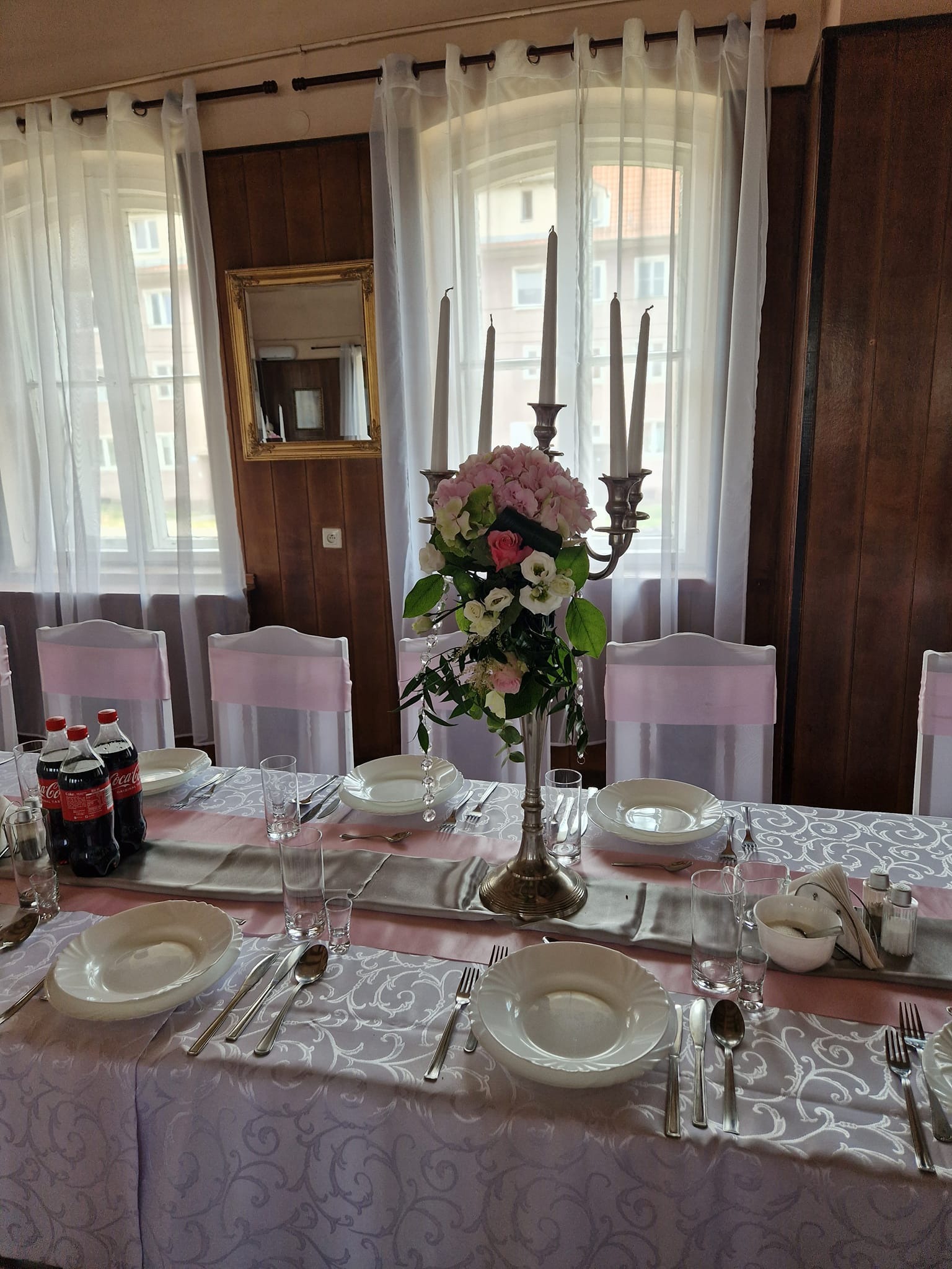 Elegancko nakryty stół z białymi talerzami, sztućcami, szklankami, świecami i kwiatową dekoracją. Krzesła z różowymi wstążkami. Przygotowania do uroczystości.