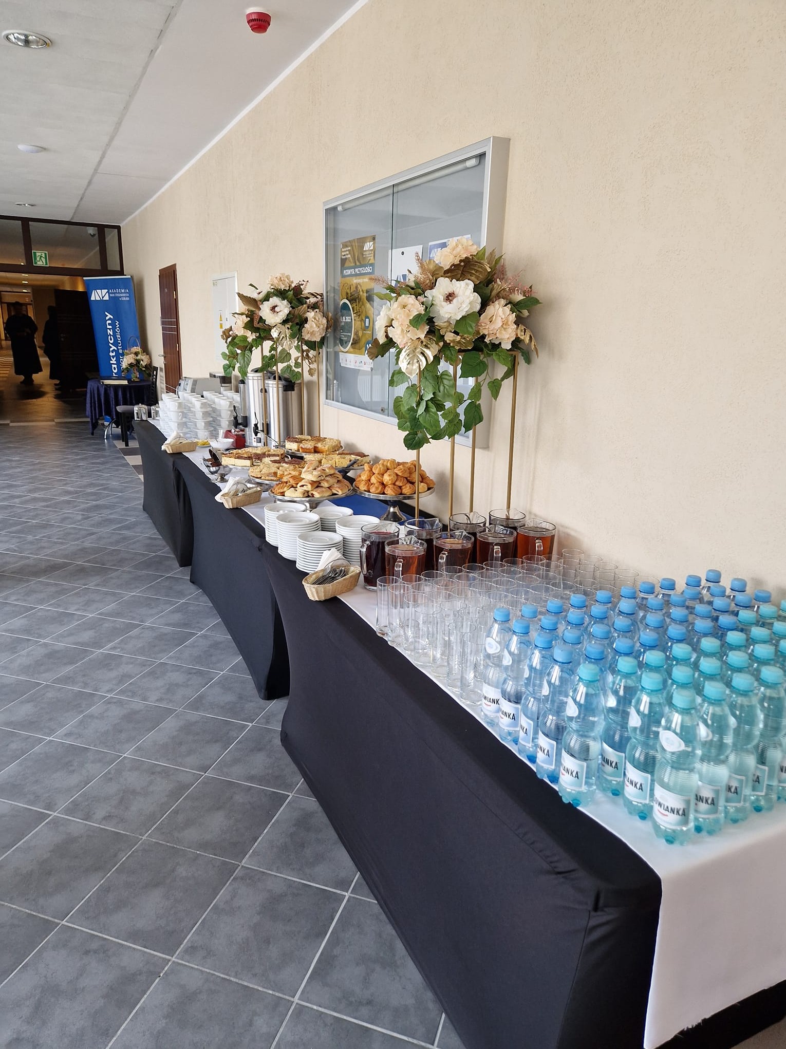 Stół cateringowy z napojami, ciastami i dekoracjami kwiatowymi w jasnym wnętrzu. Widoczne butelki wody mineralnej, szklanki, ciasta i patery.