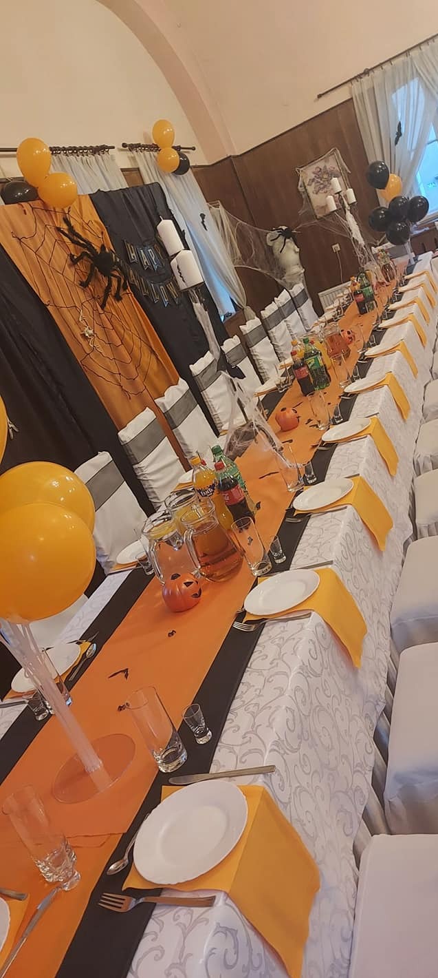 Długi stół udekorowany na Halloween: pomarańczowy bieżnik, czarne akcenty, balony, pajęczyny, dynie. Przygotowane nakrycia, napoje w dzbankach.