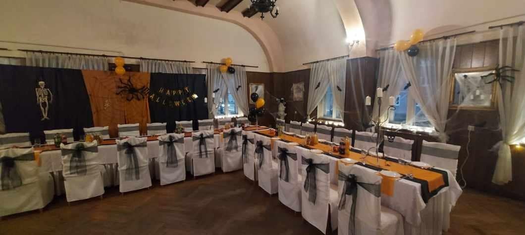 Sala bankietowa udekorowana na Halloween: długi stół z pomarańczowym bieżnikiem, krzesła z czarnymi kokardami, dekoracje z pajęczyn i balonów.
