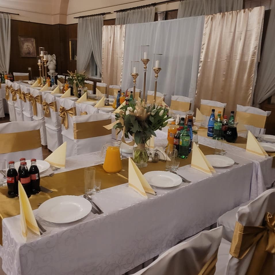 Uroczysty stół z białym obrusem, złotymi bieżnikami i dekoracjami. Krzesła z kokardami, napoje, zastawa i kwiaty. Przygotowania do imprezy.