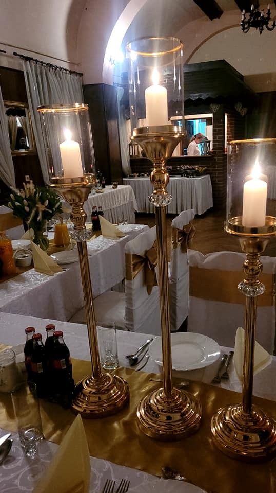 Elegancka dekoracja stołu z wysokimi, złotymi świecznikami i płonącymi świecami, ustawiona na białym obrusie z akcentami w kolorze złota. Uroczysta aranżacja.