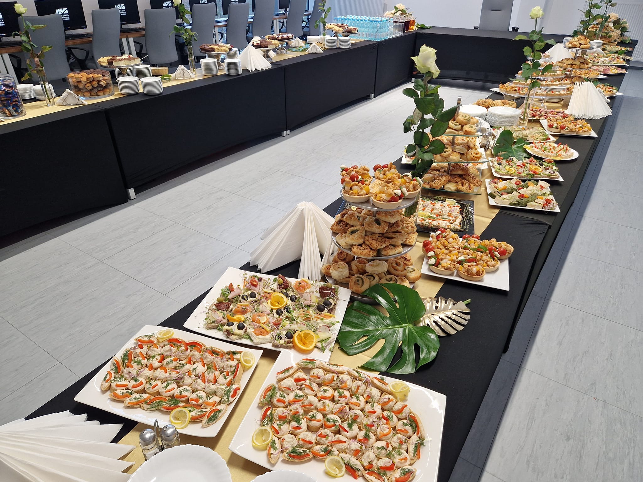 Stół z różnorodnym cateringiem: kanapki, sałatki i słodkości. Dekoracje z białych róż i tropikalnych liści. Idealne na firmowe przyjęcie.