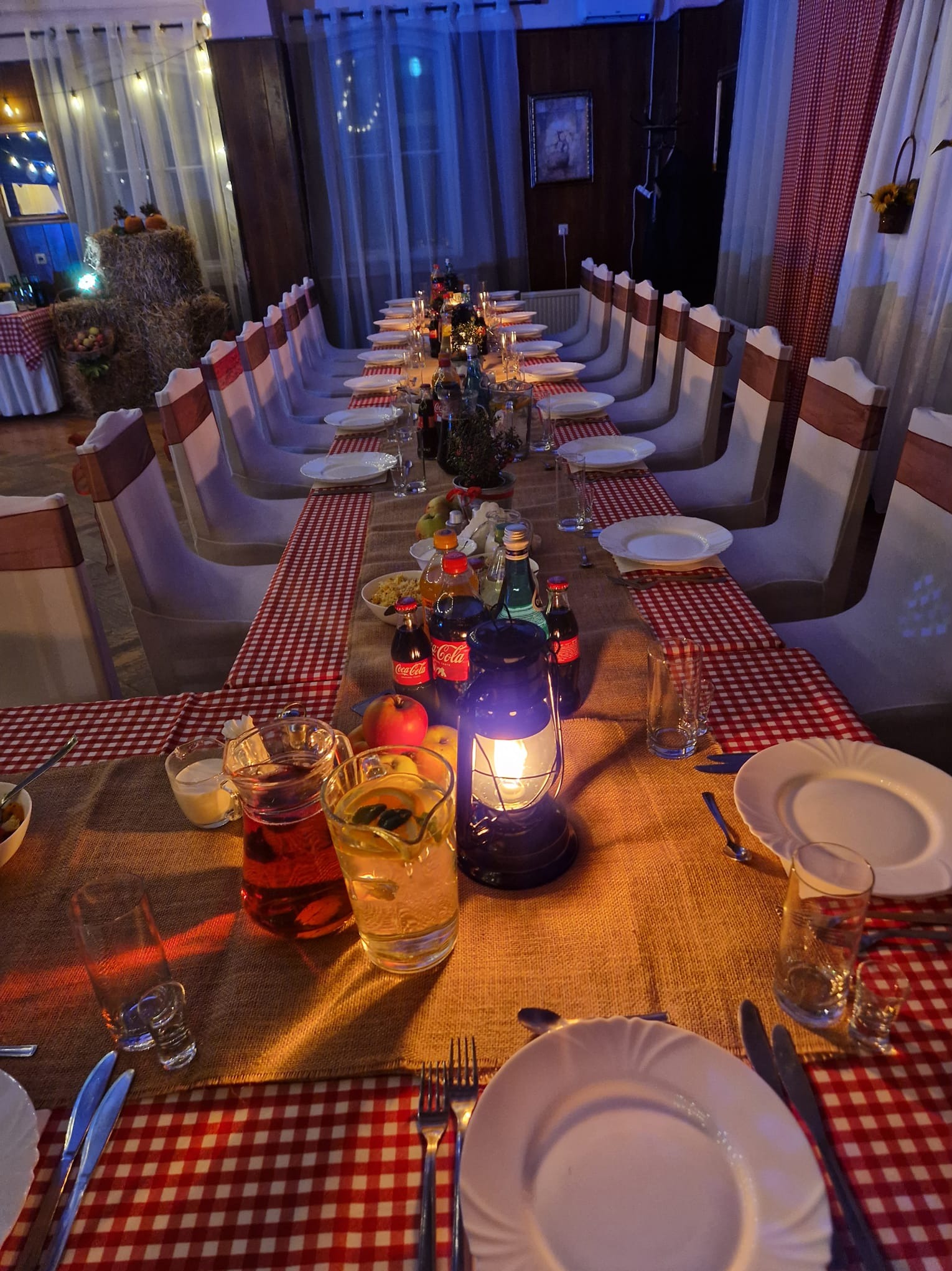 Uroczysty stół w stylu rustykalnym: lampion, napoje, zastawa, dekoracje z siana i dyni, krzesła z brązowymi szarfami. Przytulne oświetlenie tworzy ciepłą atmosferę.