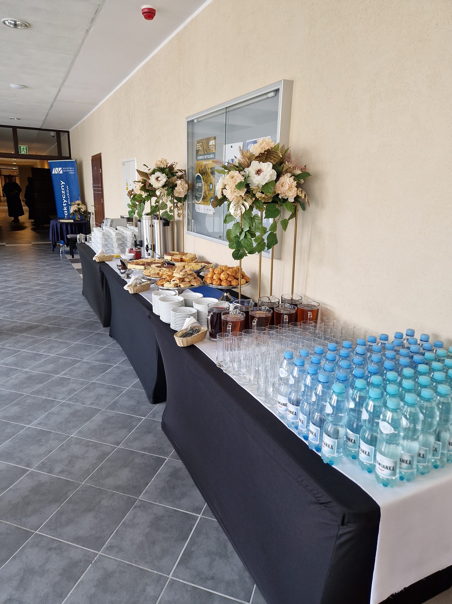 Stół cateringowy z przekąskami, napojami i kwiatowymi dekoracjami w jasnym wnętrzu, przygotowany na wydarzenie firmowe lub konferencję.
