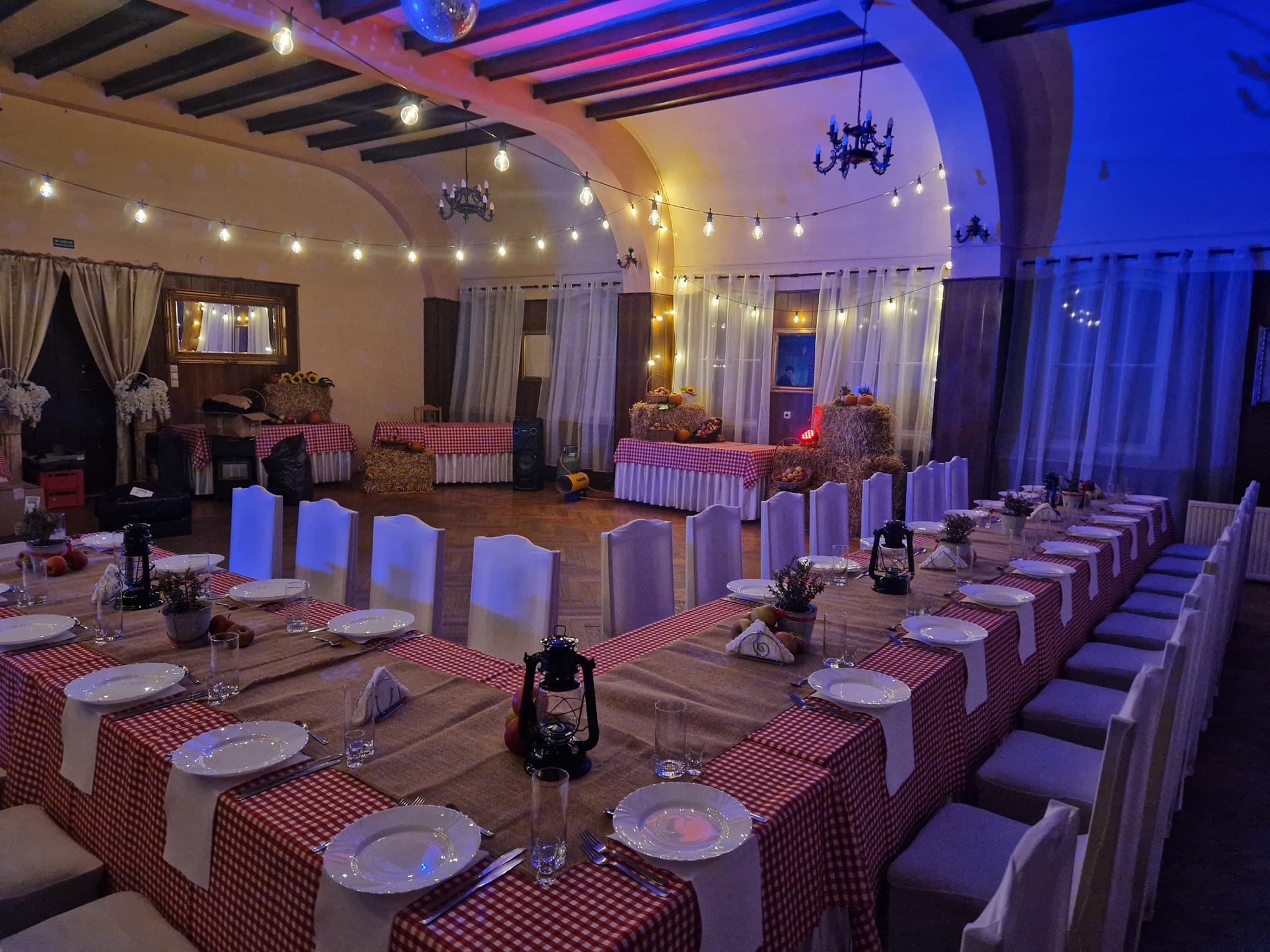 Rustykalna aranżacja sali bankietowej: długi stół z lampionami, nakrycia w czerwoną kratę, dekoracje z dyni i siana, oświetlenie girlandami i dyskotekową kulą.