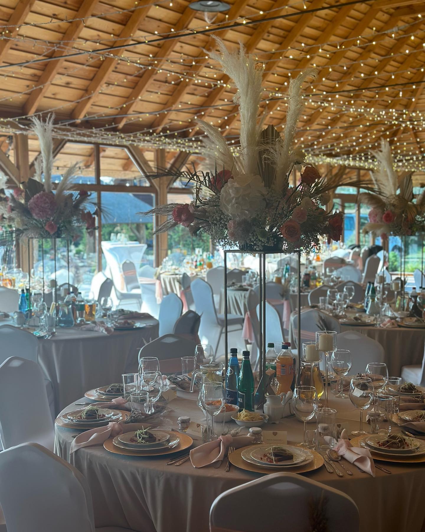 Elegancko udekorowane stoły z kwiatowymi kompozycjami i zastawą w sali weselnej z drewnianym sufitem i oświetleniem girlandami. Ustawione krzesła i nakrycia.