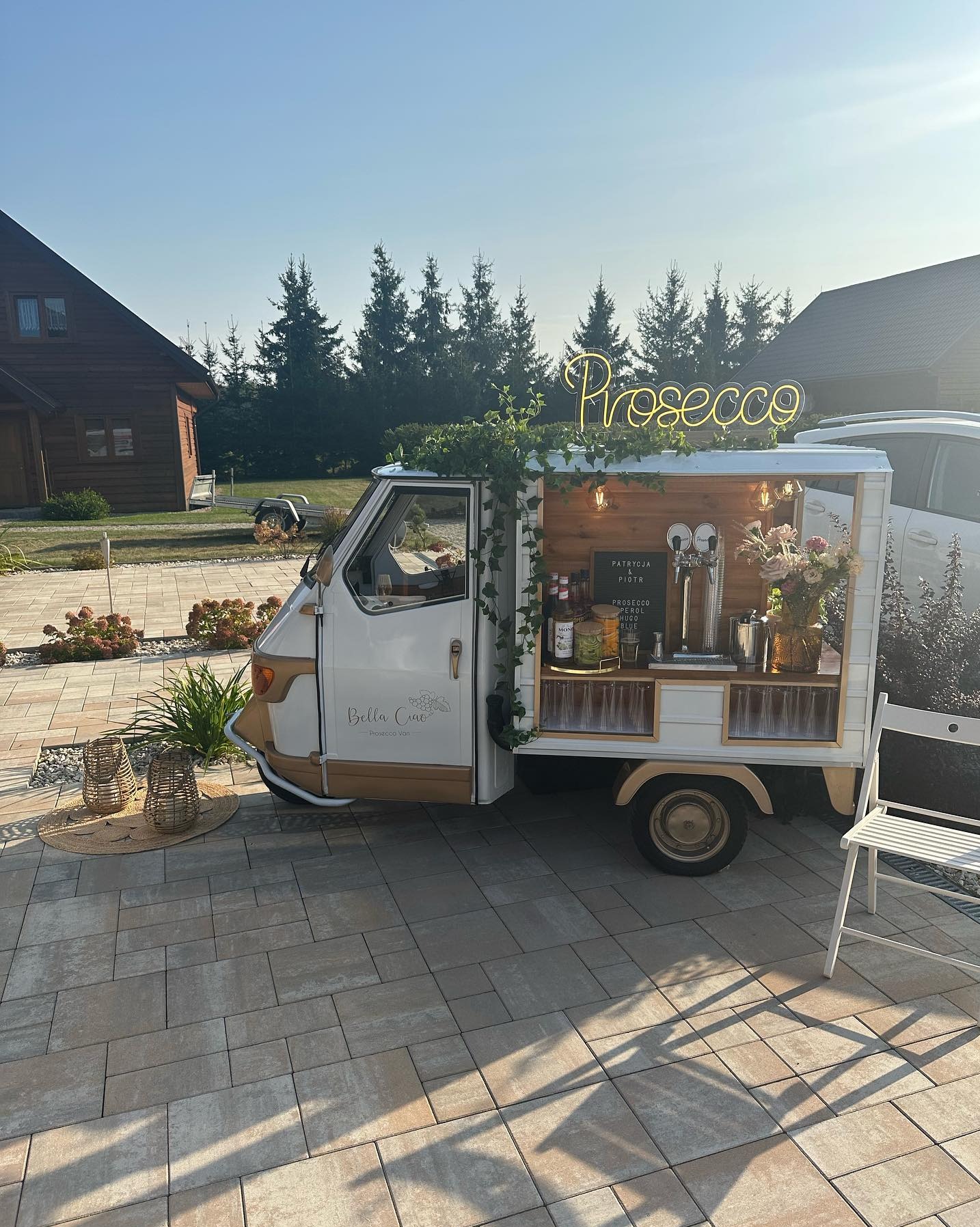 Mobilny bar prosecco 'Bella Ciao' na zewnątrz, z neonowym napisem, kwiatami i butelkami. Drewniany domek z nalewakiem i szklankami na kostce brukowej.