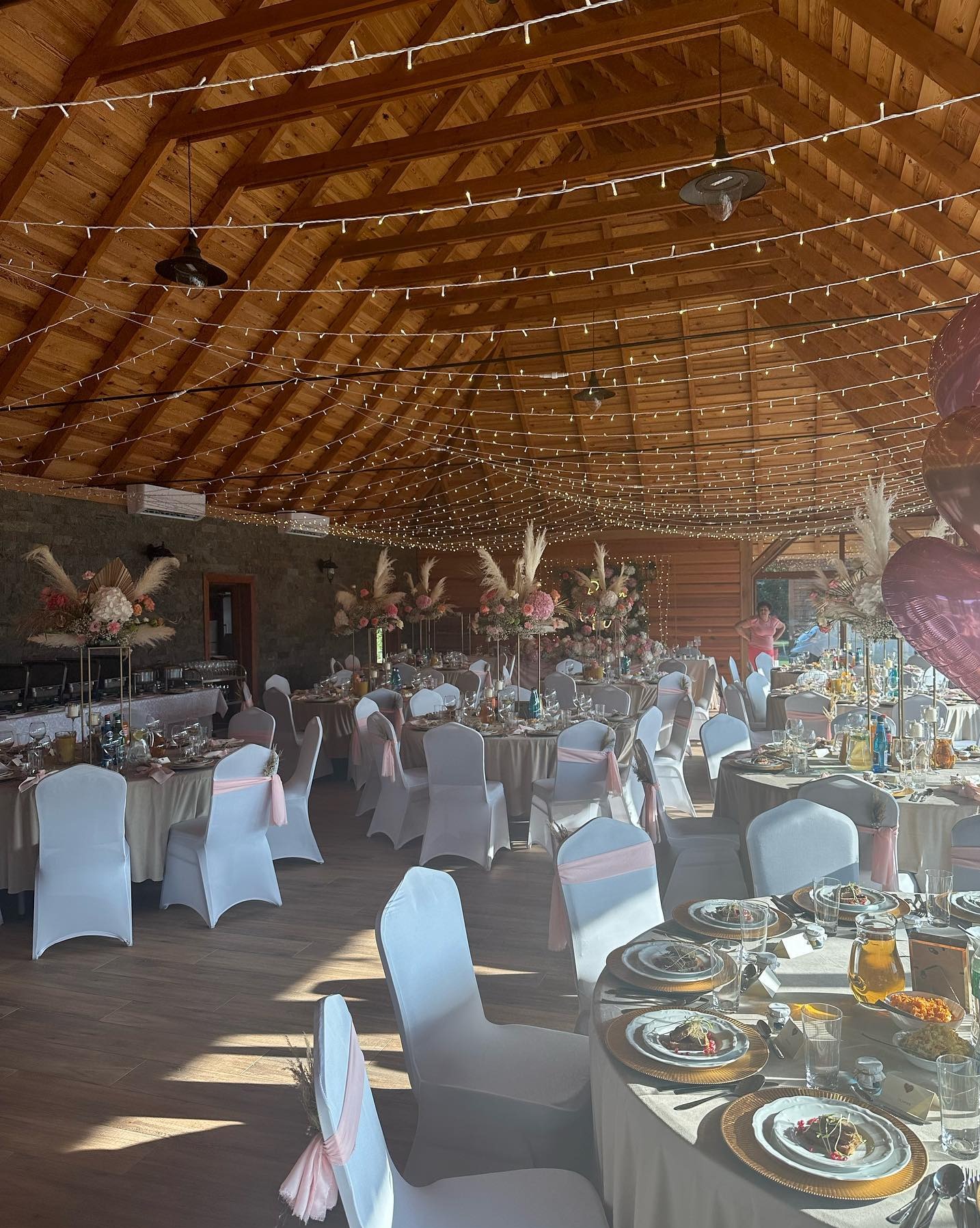 Elegancka sala bankietowa z drewnianym sufitem, udekorowana girlandami świetlnymi i kwiatowymi kompozycjami na stołach z białymi krzesłami i różowymi akcentami.