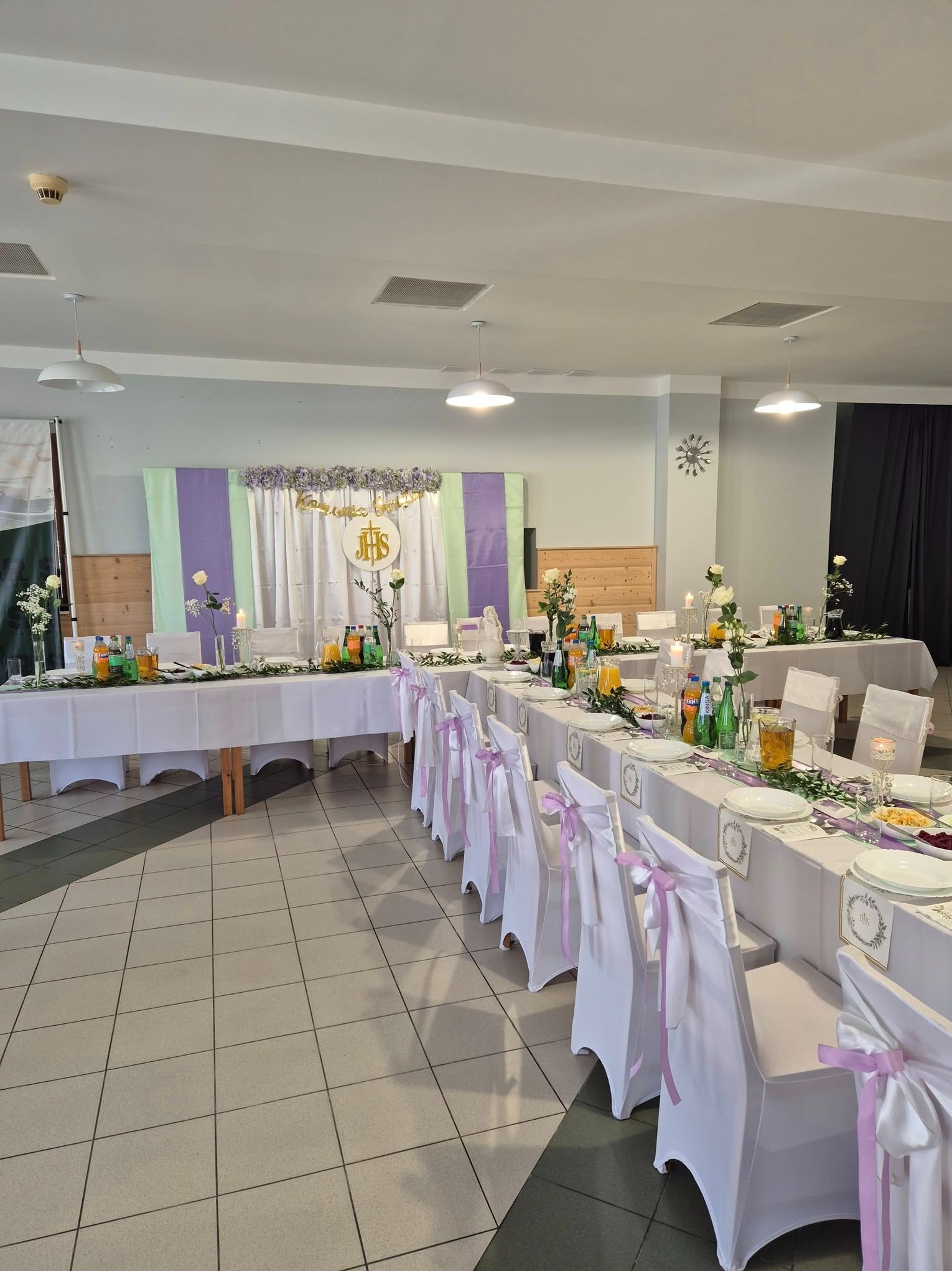 Uroczysty stół komunijny z dekoracją kwiatową, białymi obrusami i krzesłami z fioletowymi wstążkami. Napoje i dania na stole. Pastelowe tło z napisem IHS.