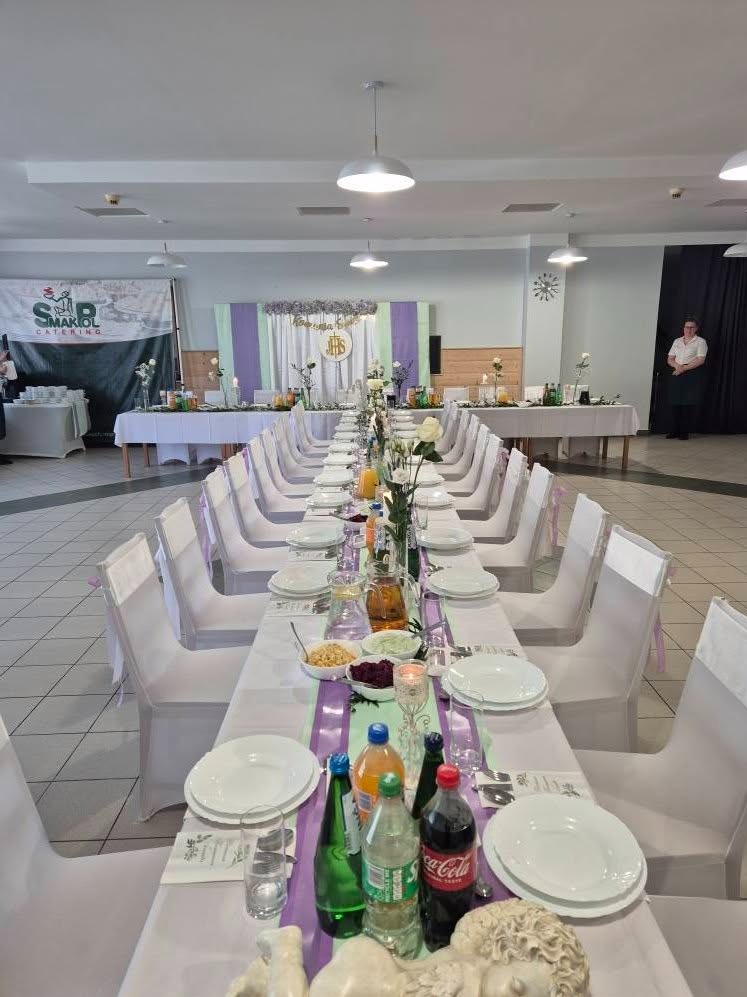 Uroczysty, długi stół z białymi krzesłami, nakryty do przyjęcia. Dekoracje kwiatowe, napoje i zastawa. W tle baner cateringowy i pracownik w uniformie.