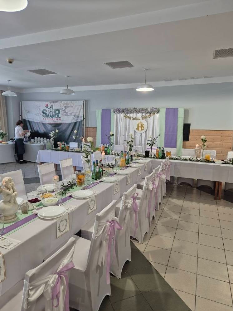 Uroczysty stół komunijny: białe obrusy, pastelowe wstążki na krzesłach, kwiaty, świece, napoje. W tle baner cateringowy i dekoracja IHS.