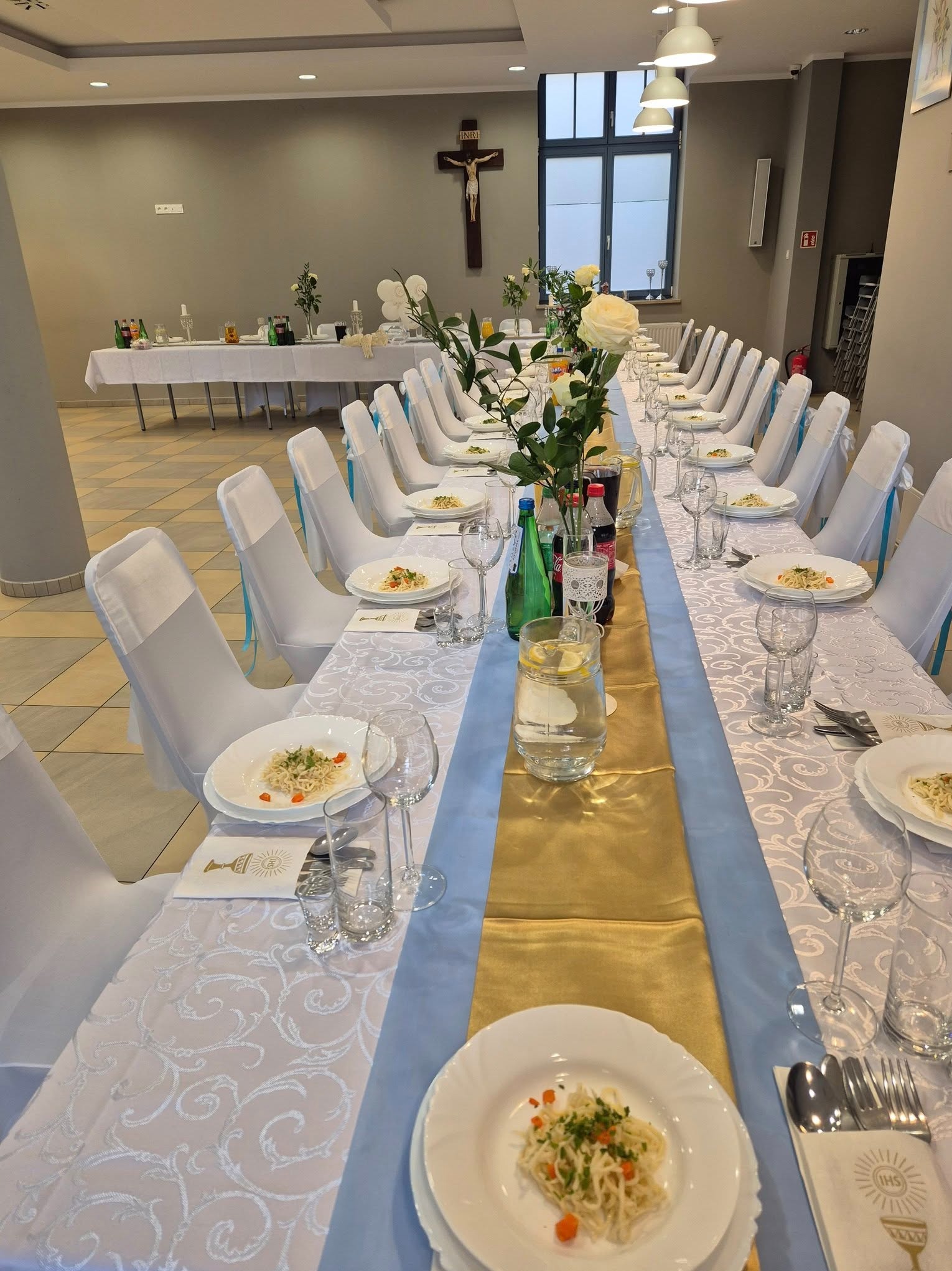 Uroczysty stół komunijny: białe pokrowce na krzesłach z błękitnymi wstążkami, złoty bieżnik, danie na talerzu, woda z cytryną, kwiaty. Krzyż na ścianie w tle.