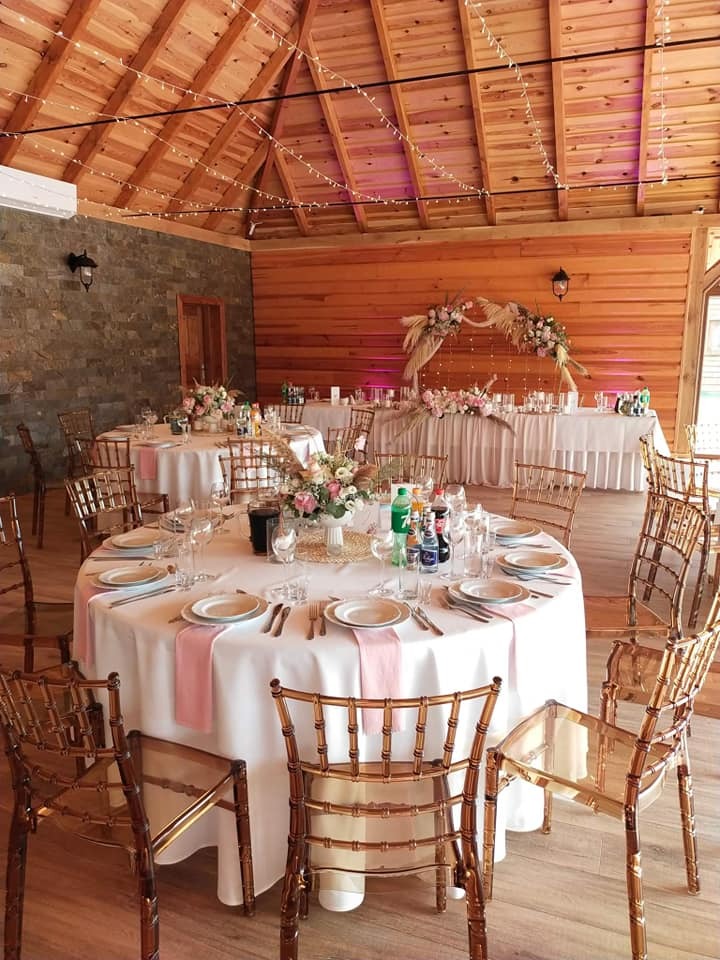 Elegancko nakryte stoły z kwiatami i dekoracjami w drewnianej sali z oświetleniem girlandami, przygotowane na przyjęcie. Transparentne krzesła.