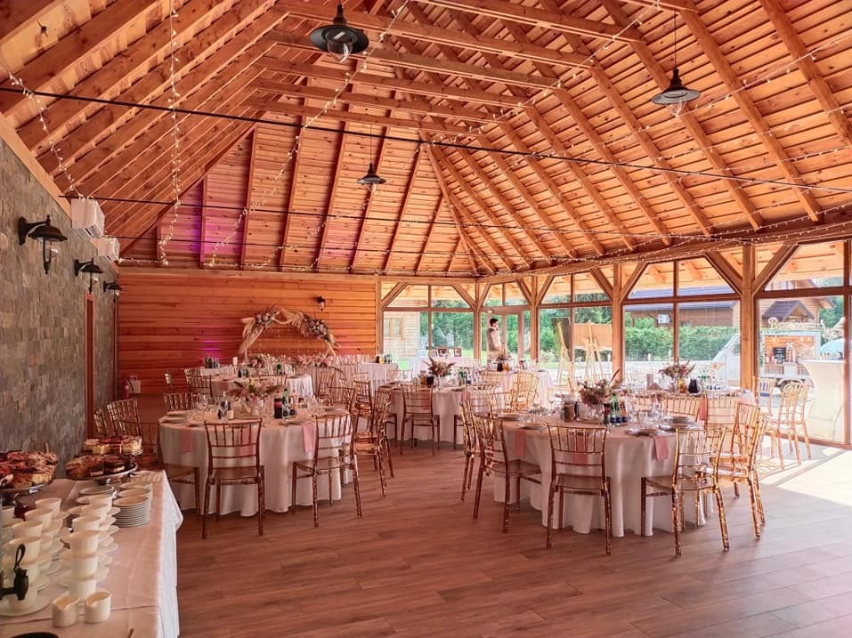 Sala bankietowa z drewnianą konstrukcją dachu, okrągłymi stołami z białymi obrusami i dekoracjami kwiatowymi, przygotowana na przyjęcie.