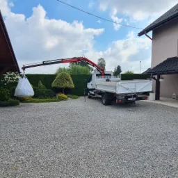 Biała ciężarówka z żurawiem podnosi duży biały worek z ogrodu na żwirowym podjeździe, widoczny dom z brązowym dachem i garaż.