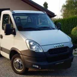 Biały samoch&oacute;d dostawczy Iveco z otwartą skrzynią ładunkową zaparkowany na żwirowym podjeździe przed domem z zadbanym ogrodem.