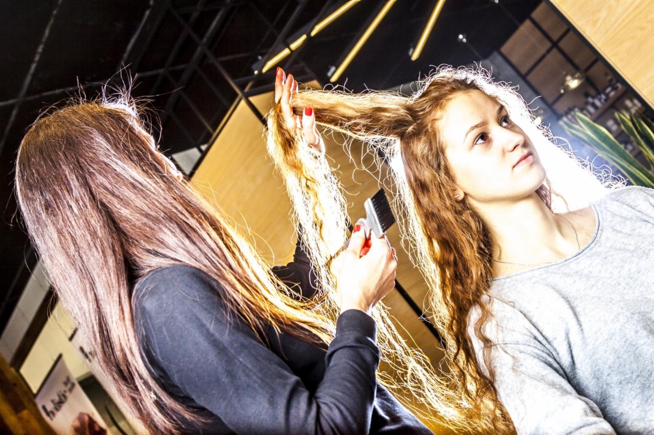 Fryzjerka z czerwonym manicure czesze długie, kręcone włosy klientki w salonie; widoczny fragment sufitu z oświetleniem.