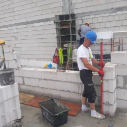Murarz w niebieskim kasku, z czerwoną poziomicą, w trakcie wznoszenia ściany z białych bloczków betonowych, widoczne szare spoiny, w tle szalunek kolumny i pracownicy w żółtych kaskach