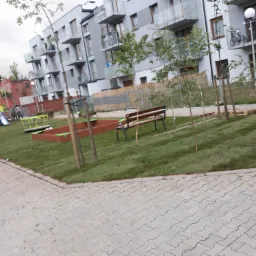 Nowo założony trawnik z młodymi drzewkami i elementami placu zabaw na tle nowoczesnego budynku mieszkalnego z balkonami, widoczne kostki brukowe na pierwszym planie.