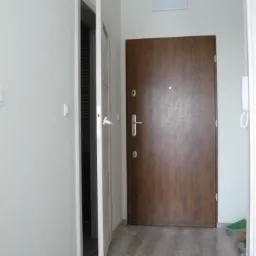 Korytarz z drewnopodobną podłogą, białymi ścianami i dwoma drzwiami: otwartymi białymi prowadzącymi do innego pomieszczenia i zamkniętymi, brązowymi, wejściowymi.