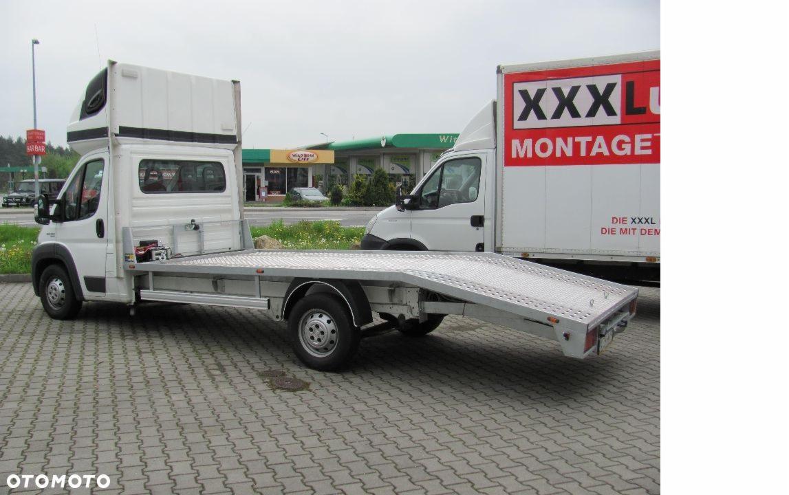 Biała laweta z aluminiową platformą, ustawiona na parkingu z kostki brukowej, w tle budynek stacji benzynowej i samochód dostawczy z reklamą.