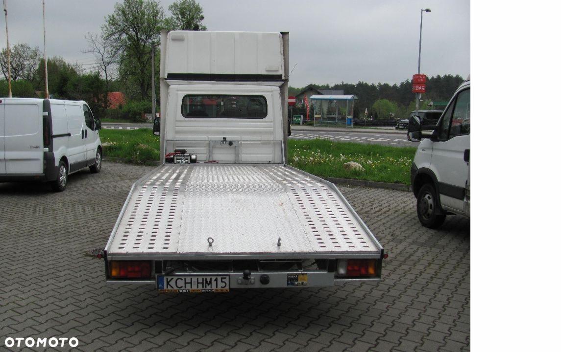 Tył białej lawety z aluminiową platformą, widoczne tablice rejestracyjne KCH HM15, w tle zaparkowane busy i otoczenie miejskie.