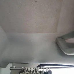 Wnętrze samochodu z widocznym sufitem tapicerowanym, fragment po czyszczeniu kontrastuje z zabrudzoną częścią, uchwyt nad drzwiami po prawej stronie, w tle wieszaki z ubraniami.